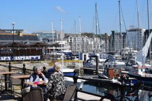 Two women sit across from each other dining outside The Harbour, in the background we can see Cap'n Jaspers restaurant and boats in the harbour.