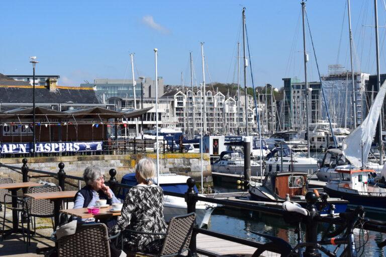 Two women sit across from each other dining outside The Harbour, in the background we can see Cap'n Jaspers restaurant and boats in the harbour.