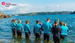 A group of people in wetsuits and snorkels stand in the water and listen to their instructors.
